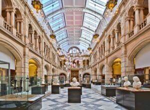 A large two-storey atrium with a glass roof, stone neo-classical pillars, and ornate chandeliers. Scattered throughout the room are busts and small statues on metal plinths.