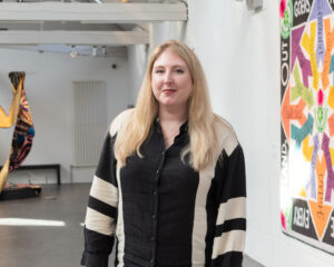 An adult with light skin and long blonde hair, wearing a cream and black shirt.