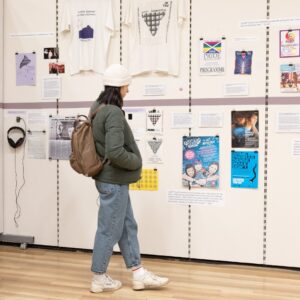 An adult with light skin, white hat, and green jacket views posters, t-shirts, and photos relating to LGBTQIA+ life in Scotland on display at the Living Memory Association in Leith.