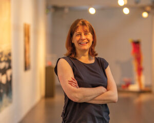 A light skinned person with shoulder length, straight auburn hair, wearing a sleeveless dark grey top, stands with their arms crossed