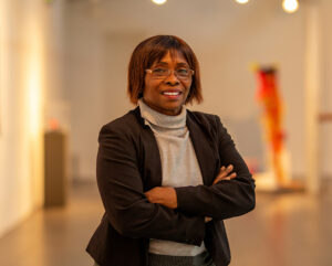 A dark skinned person with chin length bobbed straight black hair, wearing a light grey roll neck top and an black jacket, stands with their arms crossed