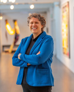 A light skinned person with short, cropped, curly grey hair, wearing a black blouse and a petrol blue blazer with her arms crossed over her chest