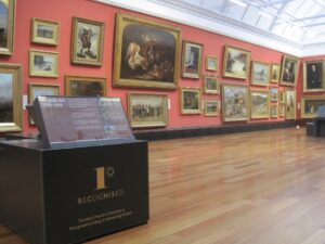 A collection of large 19th centuries paintings displayed on a red wall at the McManus Gallery. In the foreground is a sign promoting the artworks as a Recognised Collection.