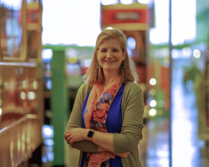 A light skinned person with shoulder length straight blonde hair, wearing a blue dress, green cardigan and a floral scarf stands with their arms crossed