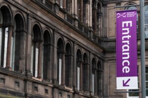 A tall purple entrance sign at the National Museum of Scotland in Edinburgh. It is pointing towards a building with long rows of ornate stone arches.