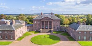 A large neoclassical house made of pale red stone. The house has two large wings and a courtyard with a lawn.