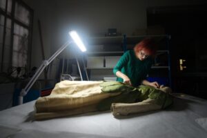 An adult who has light skin and red hair and is wearing a green jumper holds a measuring tape against a green silk dress. A lamp shines an intense white light onto the dress.