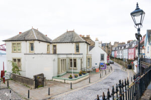 A two storey white building situated on a narrow, cobbled high street.