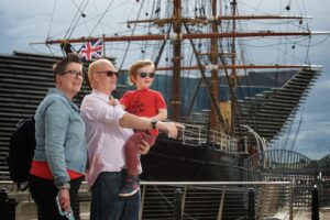 Two adults and a child with light skin smile and stand together in front of the RRS Discovery, a ship with tall wooden masts and complex rigging.