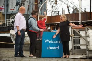 An adult with light skin, blonde hair, and glasses welcomes two adults with light skin and a child with light skin to the RRS Discovery. The child sits on a blue counter which features the words 'Welcome aboard' in white text.