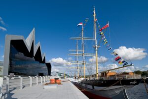 The Tall Ship Glenlee, moored next to the angular glass and metal wall of the Riverside Museum in Glasgow. The ship has three yellow masts and rows of colourful flags.
