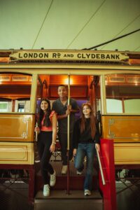 A teenager with medium-light skin and long brown hair, a teenager with medium-dark skin and short brown hair, and a teenager with light skin and long red hair stand in the doorway of a brown metal and wood tram. A sign on the side of the tram reads 'London Road and Clydebank'.