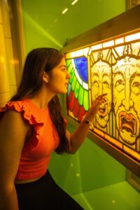 A teenager with medium-light skin and long brown hair raises their hand up to a stained glass panel. The panel is decorated with two detailed faces.