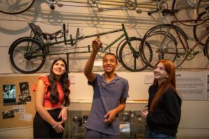 A teenager with medium-light skin and long brown hair, a teenager with medium-dark skin and short brown hair, and a teenager with light skin and long red hair stand in front of a display of bicycles. The teenager with medium-dark skin points up at the ceiling and smiles.