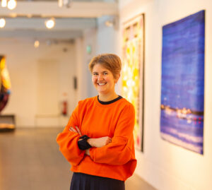 A light skinned person with short bobbed blonde hair, wearing a bright orange jumper, stands with their arms crossed