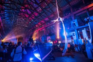 A rope dancer performs in a large industrial space as an audience watches from below. The space is bathed in pink and blue light.
