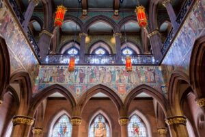 A foyer with ornately painted murals, rows of carved stone arches, and stained glass windows. Gold and red lamps hang from the ceiling.