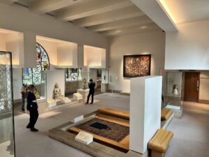 The interior of a museum gallery with glass cases displaying religious sculptures and artifacts. A large ornate rug is shown on a raised wooden platform surrounded by curved benches. Colourful arched stained glass windows line the walls, and a large framed painting hangs on the back wall. Visitors observe the objects. The space features white walls, recessed ceiling lights, and informational plaques.