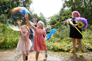 Two young children with light skin and pink dresses reach out to grab a large bubble in a garden. An adult with light skin, pink hair, and a black dress looks on and smiles.