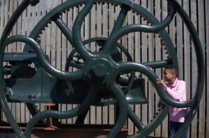 A child with medium-dark skin and a pink t-shirt smiles while standing between the large dark green metal wheels of an industrial machine.