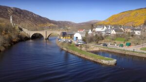 The village of Helmsdale. Situated next to a stone bridge which crosses a river is Timespan Heritage Centre, a metal building with grey and yellow walls.