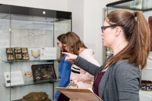 An adult with light skin, brown hair, and glasses holds a clipboard and points at a museum case filled with biological specimens. Two adults with light skin, brown hair, and glasses stand behind the first adult, looking into the case.