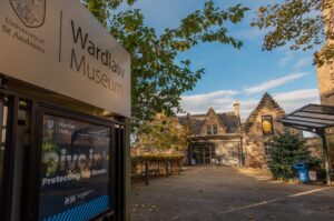 The branches of a tree hang over a sign advertising Wardlaw Museum in St Andrews. The museum, a stone building with crow-step gables, is in the background.