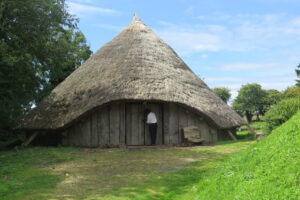 An adult wearing a white shirt and black trousers stands at the door of a wooden roundhouse with a thatched roof.
