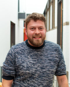 An adult with light skin, short wavy brown hair, a brown beard, and a dark blue jumper.