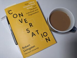 A mug of coffee and a yellow book with the title 'The Conversation' on the cover.