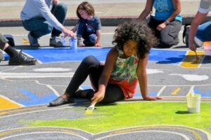 A young person with medium dark skin tone and medium length dark curly hair, sits on a school playground colouring the ground in front of them in green chalk.