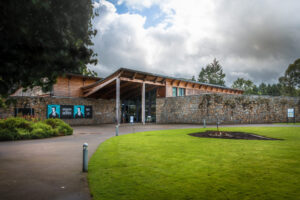 A modern building with rough stone walls and a sweeping wooden roof. A curving path leads up to the entrance.