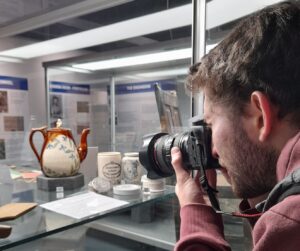 A light skinned person with short brown hair and a beard holds a camera up to their face to photograph a ceramic teapot in a glass case