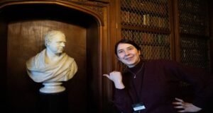 An adult with light skin and black hair smiles and gestures with their thumb towards a white bust of Sir Walter Scott, an 18th century Scottish author.