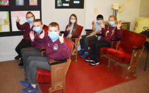 A group of children all wearing maroon jumpers and grey or black trousers and face masks are sitting in red and gold old fashioned cinema style seating next to colour drawings and paintings.