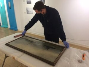 An adult wearing a face mask and plastic gloves is placing a painting of sea and boats onto a plastic covered table.