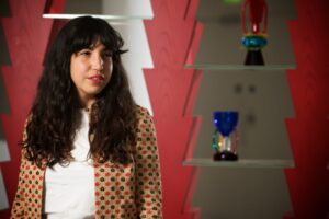 An adult with medium-light skin and long brown hair looks away to their left. In the background is a red shelving unit which displays glass objects.