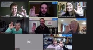 A screenshot of seven adults meeting online. Each adult is smiling at their screen and crossing their fingers.