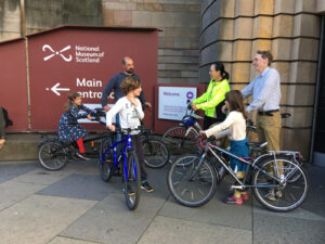 A group of adults and children stand with bicycles and talk while standing in front of the entrance to the National Museum of Scotland.