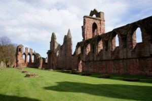 The ruins of a medieval building made of red stone. It has rows of tall arches and the remnants of a tower.