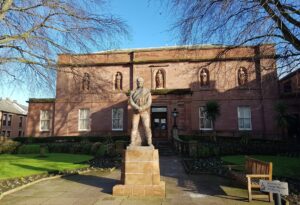 A stone statue of Robert Burns, an 18th century Scottish poet, stands on a stone plinth in front of a wide two-storey building with red stone walls and a low roof.