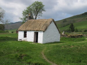 An old white stone thatched cottage surrounded by green grass and a dry stone wall.