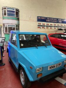A young adult with light skin and brown hair sits inside a small blue car at Dundee Museum of Transport.