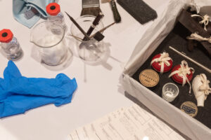Equipment used for adding accession numbers to museum objects. This includes a jar holding brushes, pens, and scalpels; a cloth; latex gloves; large brushes; and a small glass bottle of clear fluid.