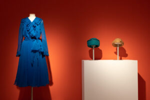 A blue gown and two fabric hats on display in front of a bright orange wall.