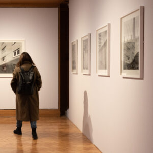 An adult with long brown hair and a dark green coat walks past a row of black and white photos on display in a gallery.