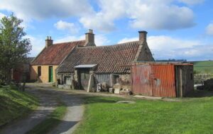 A line of three small single-storey buildings in a rural setting. The buildings on the left and in the centre have red tiled rooves, while the building on the right has a flat roof and a wide double door of rusty corrugated metal.