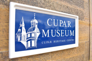 A white and blue wall-mounted sign which features the words 'Cupar Museum, Cupar Heritage Centre' and an illustration of a domed building.