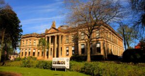 A two-storey neo-classical stone building with tall pillars and an ornate pediment. A stone statue of a human figure sits above the pediment.