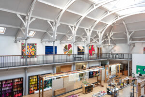 The gallery at Dovecot Studios. Colourful tapestries are displayed along the wall of a wide balcony. In the hall below are rows of weaving looms and shelves of colourful wool. Long white arches reach above the balcony, supporting a high white ceiling.